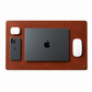 A brown leather desk mat with a small embossed logo in the bottom right corner, styled with a black laptop, a black smartphone, a white earbud case, and a white wireless mouse on a white background.